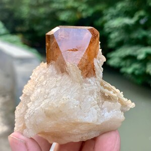 May include: A faceted, amber-colored crystal formation atop a rough, off-white mineral base. The crystal has a geometric shape with multiple facets, reflecting light. The base has a textured, layered appearance, with a natural, earthy aesthetic.