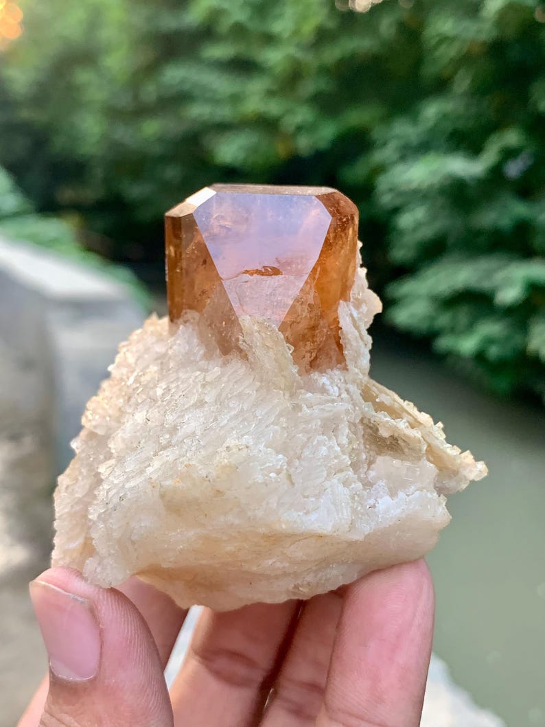 May include: A faceted, amber-colored crystal formation atop a rough, off-white mineral base. The crystal has a geometric shape with multiple facets, reflecting light. The base has a textured, layered appearance, with a natural, earthy aesthetic.