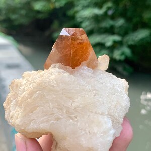 May include: A natural crystal formation featuring a large, orange-brown crystal point atop a white, textured mineral base. The crystal is held in a hand, with a blurred green background.