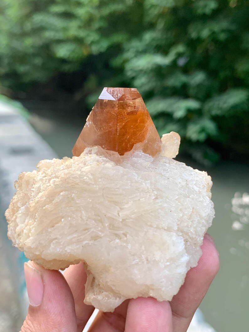 May include: A natural crystal formation featuring a large, orange-brown crystal point atop a white, textured mineral base. The crystal is held in a hand, with a blurred green background.