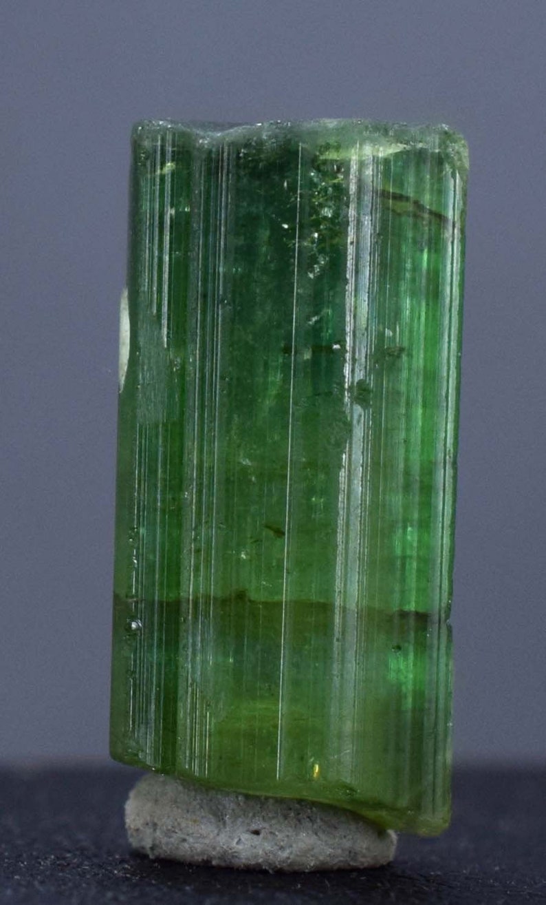 Op de afbeelding: Een levendig groene toermalijnkristal, verticaal geori&euml;nteerd, met een gestreepte textuur. De edelsteen is gemonteerd op een kleine, ruwe, grijze basis. De kleur van het kristal varieert van licht- tot diepgroen, met zichtbare interne structuren.