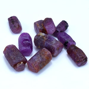 Pode incluir: Um grupo de gemas brutas roxas, provavelmente safiras, com uma variedade de formas e tamanhos. As pedras t&ecirc;m um tom roxo intenso e est&atilde;o dispostas sobre um fundo branco.