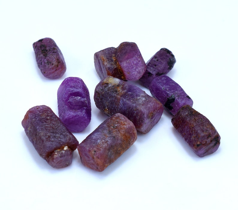 Pode incluir: Um grupo de gemas brutas roxas, provavelmente safiras, com uma variedade de formas e tamanhos. As pedras t&ecirc;m um tom roxo intenso e est&atilde;o dispostas sobre um fundo branco.