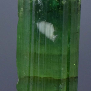 Op de afbeelding: Een doorschijnend, groene toermalijnkristal met verticale strepen en een witte insluitsel. De edelsteen is gemonteerd op een kleine, grijze, getextureerde basis. De kleur van het kristal varieert van licht- tot donkergroen.
