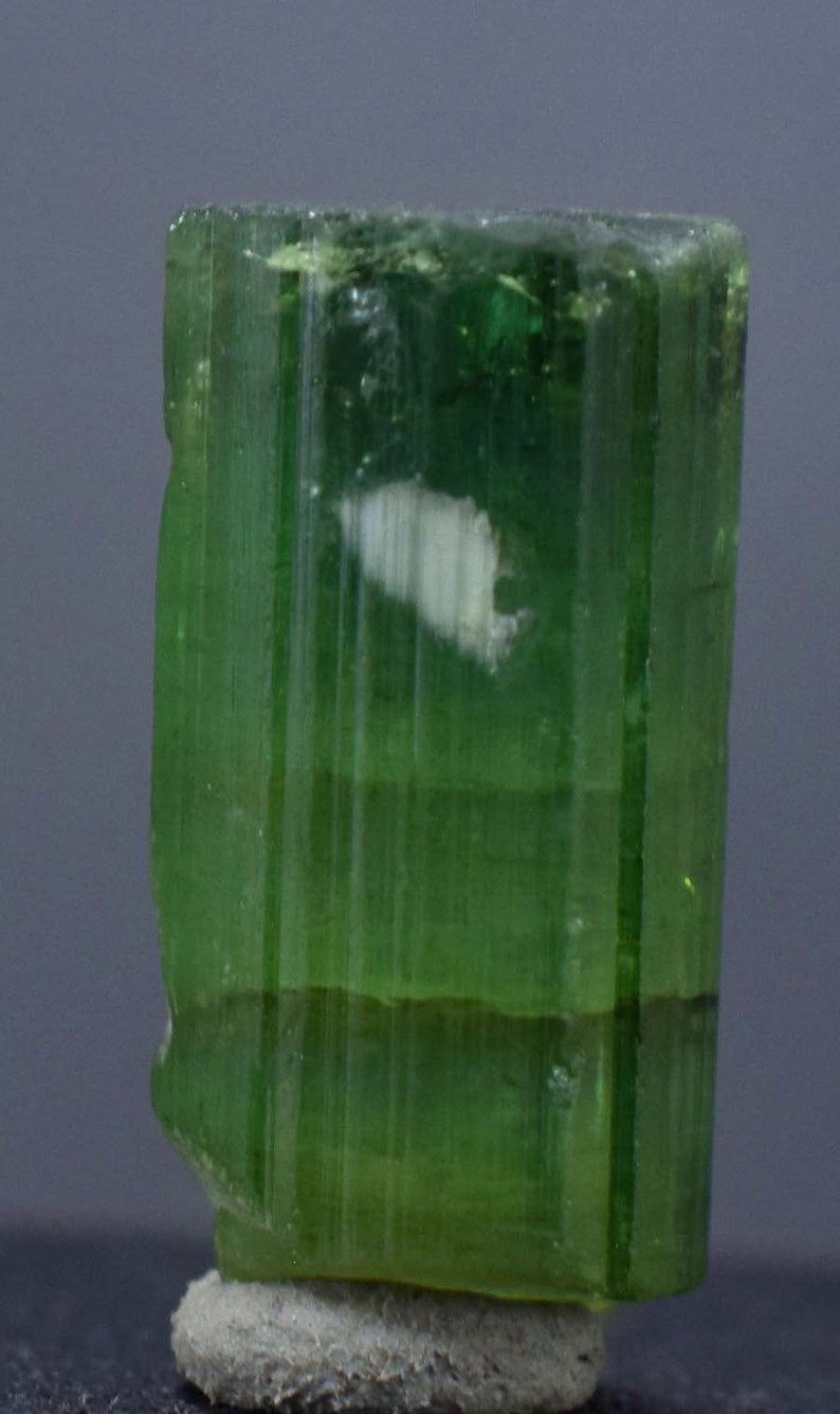 Op de afbeelding: Een doorschijnend, groene toermalijnkristal met verticale strepen en een witte insluitsel. De edelsteen is gemonteerd op een kleine, grijze, getextureerde basis. De kleur van het kristal varieert van licht- tot donkergroen.