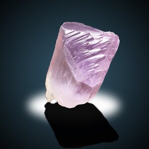 May include: A large, translucent, pink crystal with striations on its surface. The crystal is sitting on a dark blue background.