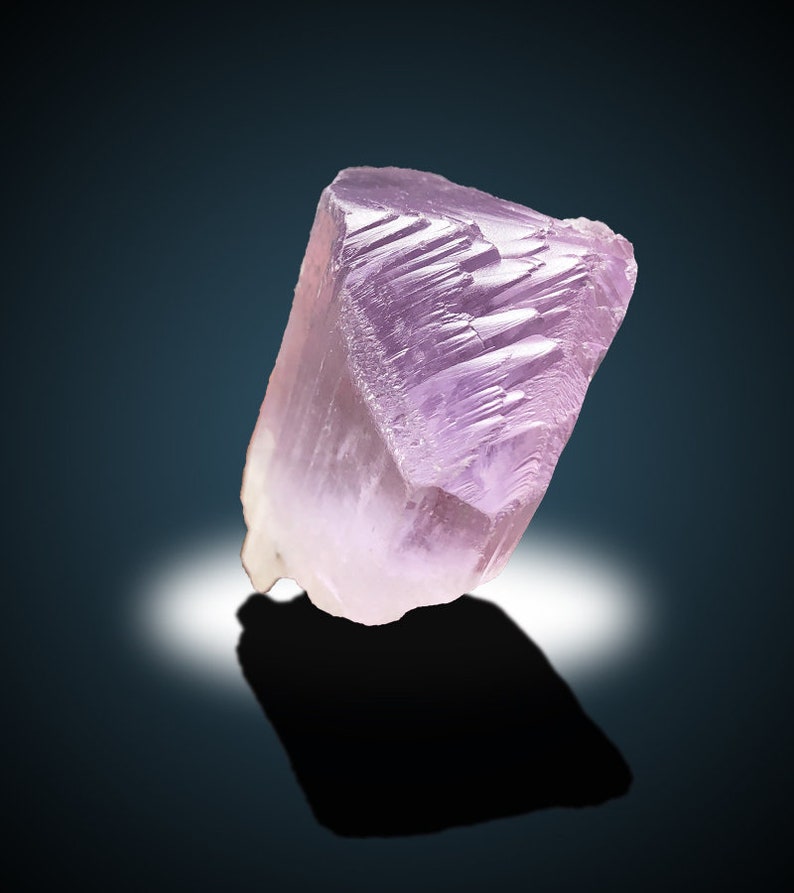 May include: A large, translucent, pink crystal with striations on its surface. The crystal is sitting on a dark blue background.