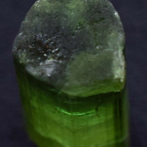 Op de afbeelding: Een close-up van een groene toermalijnkristal. De bovenkant van het kristal is een donkerder, ondoorzichtig groen, terwijl de onderkant een levendig, doorschijnend groen is. Het kristal heeft een ruw, getextureerd oppervlak.