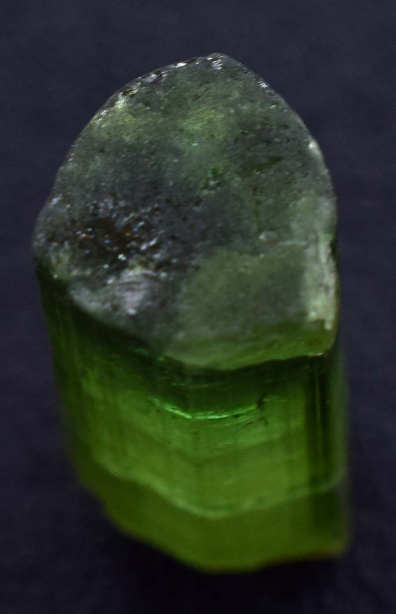 Op de afbeelding: Een close-up van een groene toermalijnkristal. De bovenkant van het kristal is een donkerder, ondoorzichtig groen, terwijl de onderkant een levendig, doorschijnend groen is. Het kristal heeft een ruw, getextureerd oppervlak.