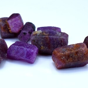 Pode incluir: Um grupo de oito gemas brutas, roxas e marrom-avermelhadas. As gemas s&atilde;o de v&aacute;rios tamanhos e formas, e t&ecirc;m um aspecto natural n&atilde;o polido.