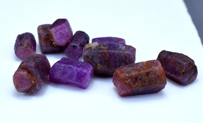 Pode incluir: Um grupo de oito gemas brutas, roxas e marrom-avermelhadas. As gemas s&atilde;o de v&aacute;rios tamanhos e formas, e t&ecirc;m um aspecto natural n&atilde;o polido.