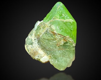 Puede incluir: Un cristal verde translúcido facetado con una textura rugosa y natural. El cristal tiene una parte superior puntiaguda y una base plana. Es un espécimen natural de peridoto verde.