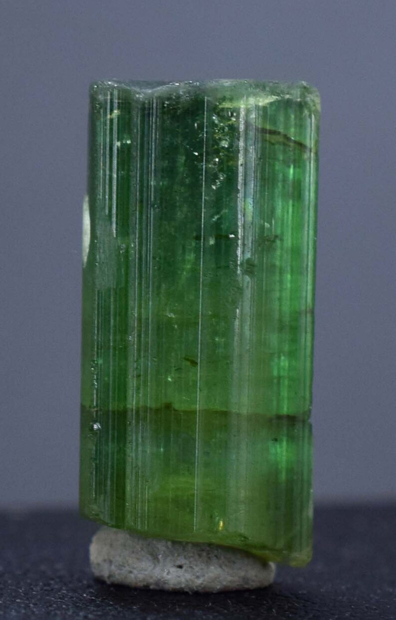 Op de afbeelding: Een doorschijnend, groene toermalijnkristal met verticale strepen. De edelsteen heeft een cilindrische vorm en is gemonteerd op een kleine, grijze basis. De kleur van het kristal varieert van licht- tot donkergroen.