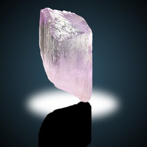 May include: A large, translucent, pale pink crystal with a geometric shape. The crystal is sitting on a dark surface with a white light reflecting off of it.