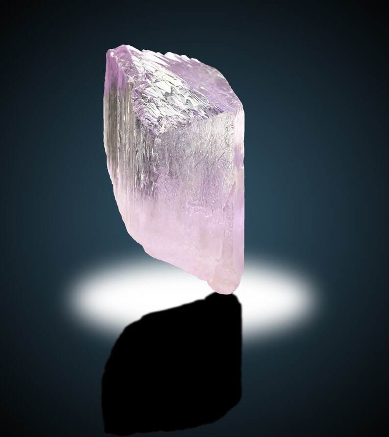 May include: A large, translucent, pale pink crystal with a geometric shape. The crystal is sitting on a dark surface with a white light reflecting off of it.