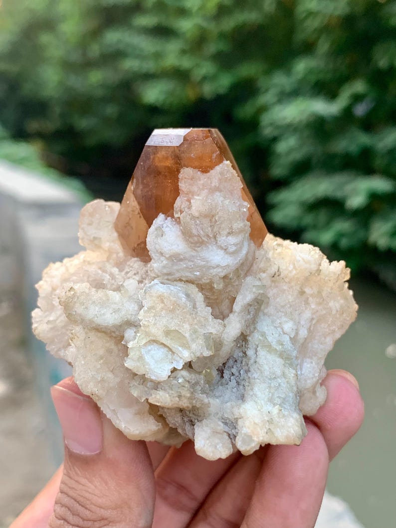 May include: A natural, raw crystal formation featuring a large, faceted, amber-colored crystal point atop a cluster of off-white, textured mineral deposits. The crystal is held in a hand, with a blurred green background.
