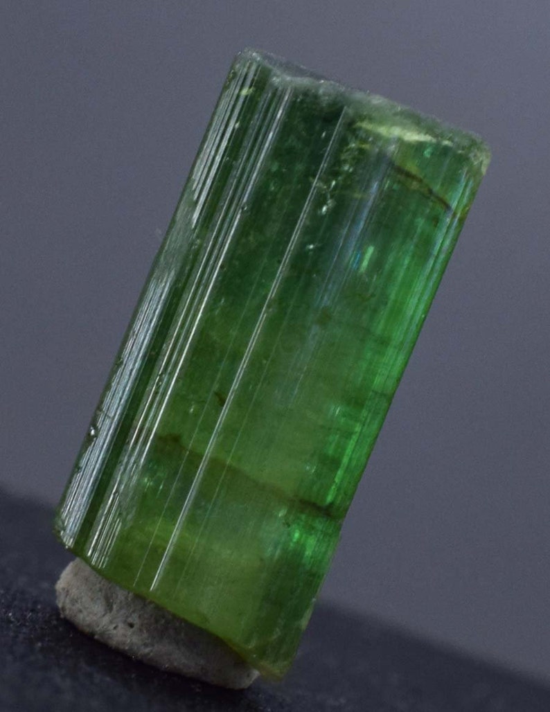 Op de afbeelding: Een levendig groene toermalijnkristal, met een rechthoekige prisma vorm met verticale strepen. De edelsteen is gemonteerd op een kleine, grijze basis, tegen een donkergrijze achtergrond. De kleur van het kristal varieert van licht- tot diepgroen.