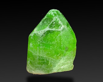 Puede incluir: Un cristal verde vibrante con una superficie rugosa y texturizada. El cristal tiene forma de pirámide con una punta puntiaguda.