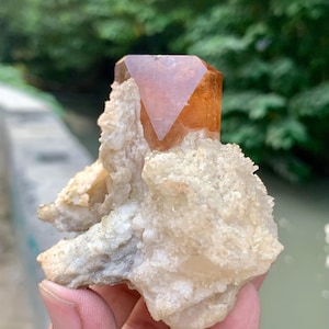 May include: A natural, raw crystal formation featuring a large, orange-brown crystal point atop a cluster of off-white, textured mineral deposits. The crystal is held in a hand, with a blurred green background.