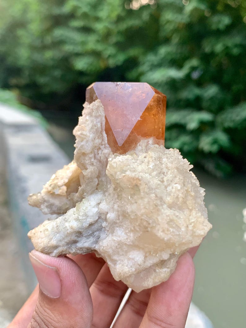 May include: A natural, raw crystal formation featuring a large, orange-brown crystal point atop a cluster of off-white, textured mineral deposits. The crystal is held in a hand, with a blurred green background.