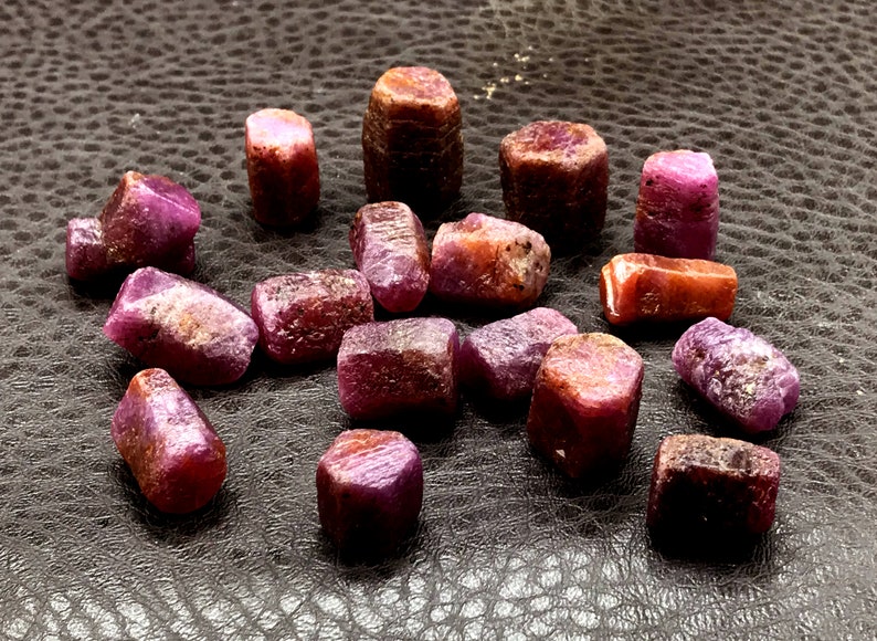 Może przedstawiać: Kolekcja 18 surowych kryształ&oacute;w rubinu, każdy o unikalnym kształcie i rozmiarze. Kryształy mają głęboki czerwony kolor z lekkim fioletowym odcieniem. Są ułożone na ciemnobrązowej, teksturowanej powierzchni.