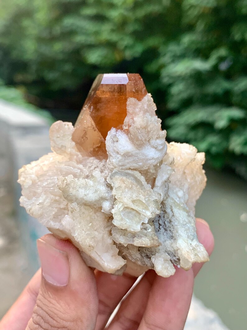 May include: A natural crystal formation featuring a large, amber-colored crystal point atop a cluster of smaller, off-white crystals. The crystal is held in a hand, with a blurred green background.
