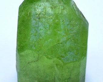 Puede incluir: Un gran cristal de peridoto verde facetado con un acabado brillante. El cristal tiene una forma ligeramente irregular y muestra algunas inclusiones internas.