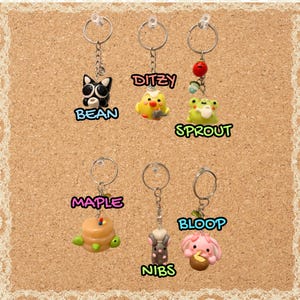 May include: Six metal keychains featuring small clay charms: a black cat labeled BEAN, a yellow chick labeled DITZY, a green frog with a red berry labeled SPROUT, a stack of pancakes labeled MAPLE, a gray mouse labeled NIBS, and a pink creature labeled BLOOP.