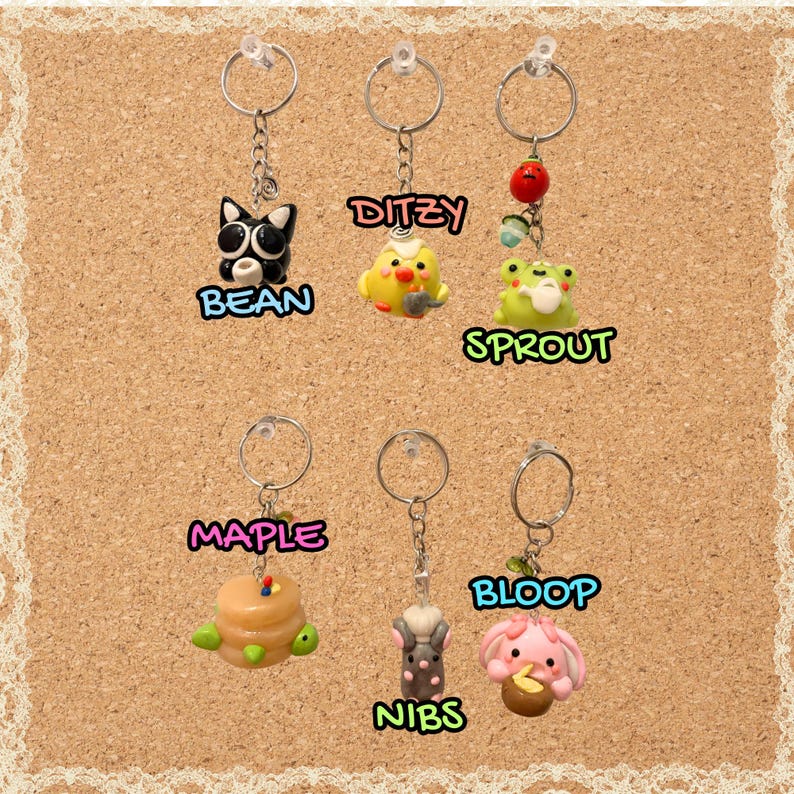 May include: Six metal keychains featuring small clay charms: a black cat labeled BEAN, a yellow chick labeled DITZY, a green frog with a red berry labeled SPROUT, a stack of pancakes labeled MAPLE, a gray mouse labeled NIBS, and a pink creature labeled BLOOP.