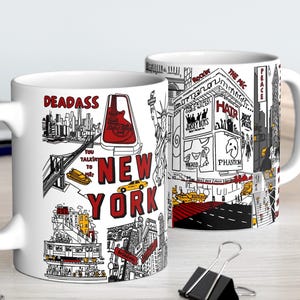 May include: Two white ceramic mugs with black and red illustrations of New York City landmarks. One mug features the text "DEADASS" and "NEW YORK". The other mug has illustrations of Times Square and other city scenes. The mugs are on a light wood surface.