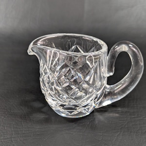 May include: A clear glass creamer with a cut crystal design. The creamer has a small handle and a rounded body.