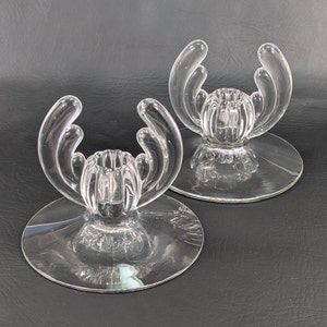 May include: Two clear glass candle holders with a unique, organic design. Each holder features a central cup for a candle and four curved arms that extend upwards.