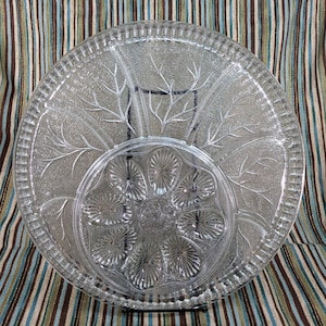 May include: A clear glass serving platter with a decorative etched design of leaves and flowers. The platter has six individual sections for serving different foods.