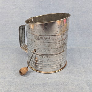 May include: A vintage silver measuring cup with a wooden handle. The cup has markings for 2, 3, 4, and 5 cups. It is a kitchen tool for baking and cooking.