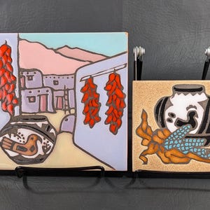 May include: Two ceramic tile wall hangings with a Southwestern theme. The first tile features a light blue, pink, and yellow scene of a pueblo village with red chili peppers hanging from a string. The second tile features a tan background with a white and black jar with a blue and orange design.