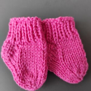 K&ouml;nnte beinhalten: Ein Paar kleine, handgestrickte Booties in leuchtendem Pink mit geripptem B&uuml;ndchen und einem einfachen glatt rechts gestrickten Muster im Fu&szlig;bereich. Die Booties sind aus dickem, strukturiertem Garn gefertigt und haben eine abgerundete Zehenform.