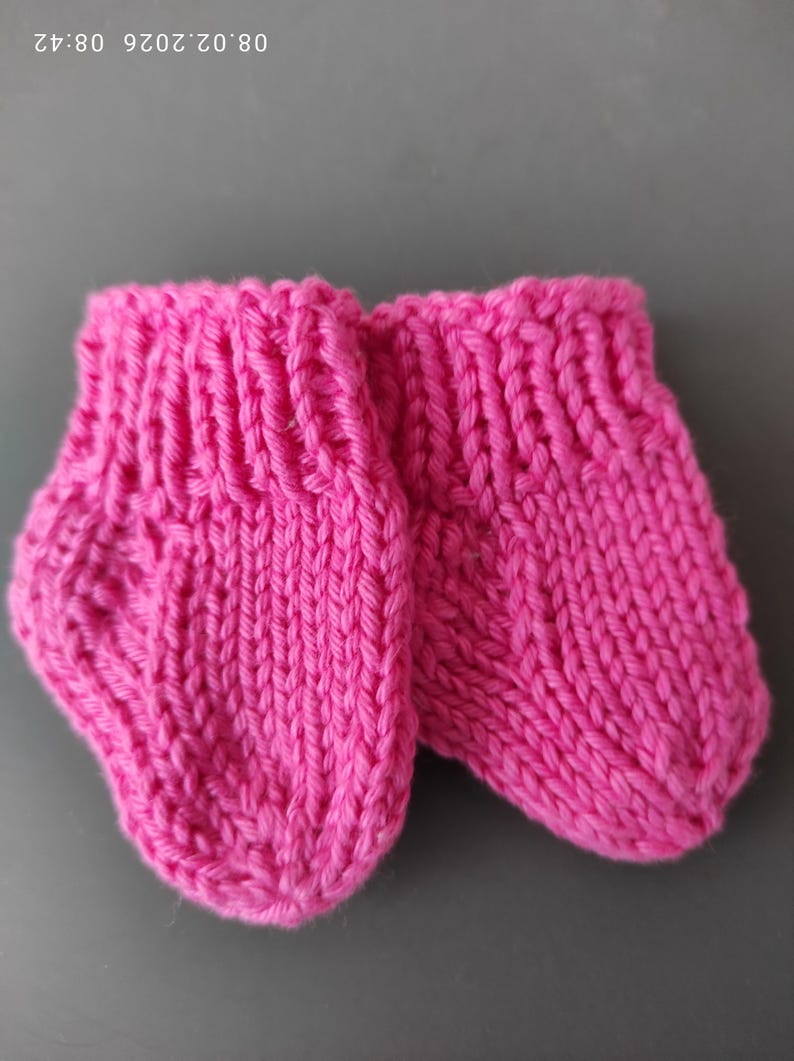 K&ouml;nnte beinhalten: Ein Paar kleine, handgestrickte Booties in leuchtendem Pink mit geripptem B&uuml;ndchen und einem einfachen glatt rechts gestrickten Muster im Fu&szlig;bereich. Die Booties sind aus dickem, strukturiertem Garn gefertigt und haben eine abgerundete Zehenform.