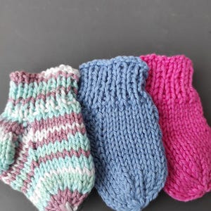 K&ouml;nnte beinhalten: Drei kleine handgestrickte F&auml;ustlinge nebeneinander angeordnet. Der erste ist gestreift in Hellblau, Wei&szlig; und Mauve. Der zweite ist einfarbig mittelblau. Der dritte ist einfarbig leuchtend pink. Jeder F&auml;ustling hat ein Rippenb&uuml;ndchen und einen Daumen.