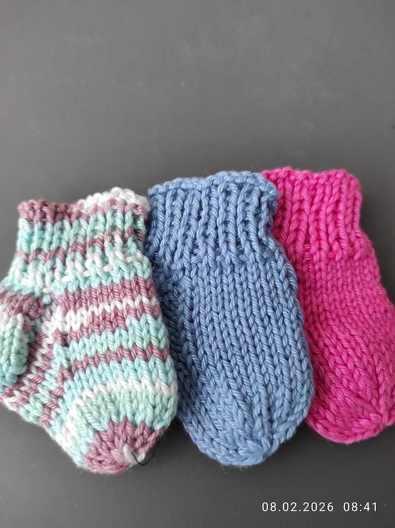 K&ouml;nnte beinhalten: Drei kleine handgestrickte F&auml;ustlinge nebeneinander angeordnet. Der erste ist gestreift in Hellblau, Wei&szlig; und Mauve. Der zweite ist einfarbig mittelblau. Der dritte ist einfarbig leuchtend pink. Jeder F&auml;ustling hat ein Rippenb&uuml;ndchen und einen Daumen.