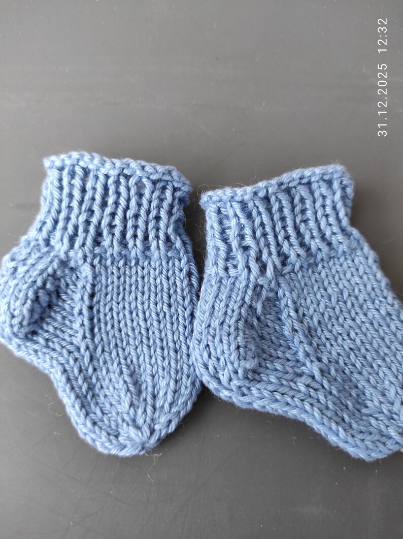 K&ouml;nnte beinhalten: Ein Paar hellblaue gestrickte Babyschuhe mit geripptem B&uuml;ndchen und einem glatt rechts gestrickten Fu&szlig;teil. Die Schuhe haben eine ausgepr&auml;gte Fersenkonstruktion und eine abgerundete Zehenpartie, gefertigt aus weichem Garn mit Struktur.