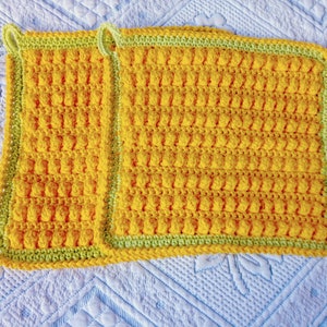Könnte beinhalten: Zwei gelbe gehäkelte Topflappen mit grünem Rand. Die Topflappen sind mit einem strukturierten Stich gearbeitet, der eine unebene Oberfläche erzeugt.
