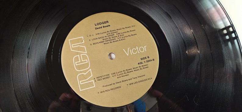 May include: A black vinyl record with a gold label featuring the album title "Lodger" by David Bowie. The label includes song titles, the RCA and Victor logos, and the text "Side B".