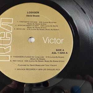 May include: A black vinyl record with a gold and white label. The label features the text "LODGER" by David Bowie, the RCA Victor logo, and a list of songs. The record is Side A, AGL 1-3254-A.