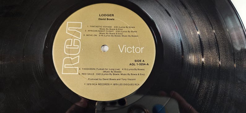 May include: A black vinyl record with a gold and white label. The label features the text "LODGER" by David Bowie, the RCA Victor logo, and a list of songs. The record is Side A, AGL 1-3254-A.