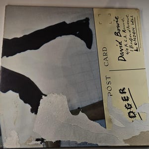 May include: A vintage postcard featuring a black and white image of a person's legs and feet. The postcard has a torn, distressed appearance with the text "POST CARD" and "David Bowie" visible. The address is listed as London, W.C.L.