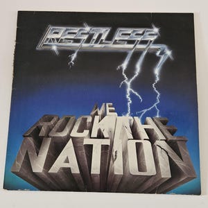 May include: Album cover for "Restless" with the band's name in a silver, blocky font, accompanied by lightning bolts. The words "We Rock the Nation" are in a 3D stone-like font against a blue and black gradient background.