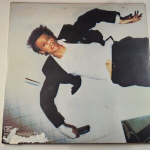 May include: Album cover featuring a man in a white shirt and black suit, seemingly falling or floating. The image has a vintage aesthetic, with a light background and a slightly worn appearance. The man is holding a gun.