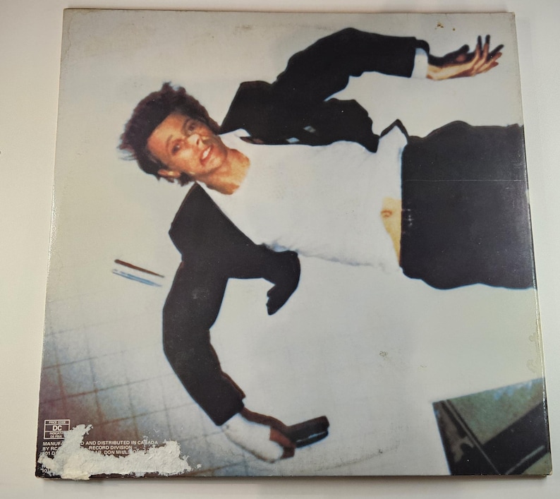 May include: Album cover featuring a man in a white shirt and black suit, seemingly falling or floating. The image has a vintage aesthetic, with a light background and a slightly worn appearance. The man is holding a gun.
