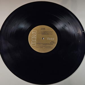 May include: A black vinyl record with a gold and white label. The label features the RCA Victor logo and the album title "Lodger" by David Bowie. The record is in good condition, with visible grooves and a glossy surface. Side A is labeled with song titles.