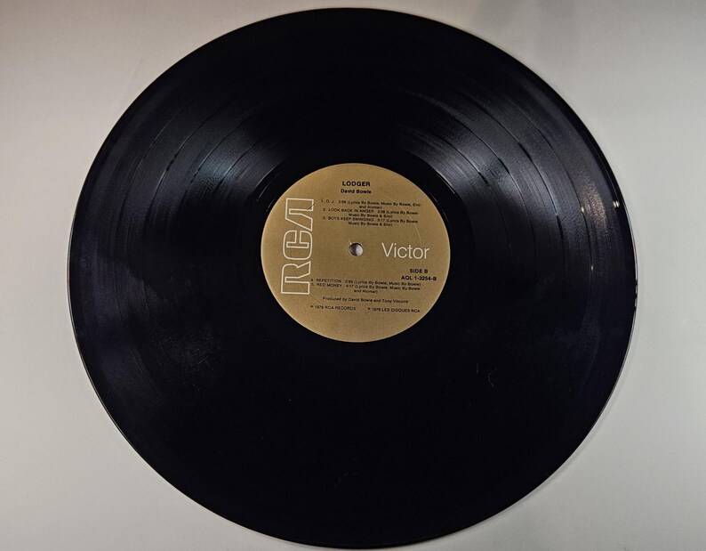 May include: A black vinyl record with a gold and white label. The label features the RCA Victor logo and the album title "Lodger" by David Bowie. The record is in good condition, with visible grooves and a glossy surface. Side A is labeled with song titles.